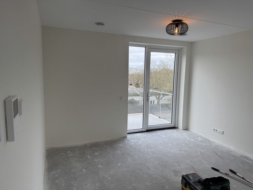 Kamer in een net opgeleverde woning met balkondeur en kale betonvloer, vlak voor het behangen